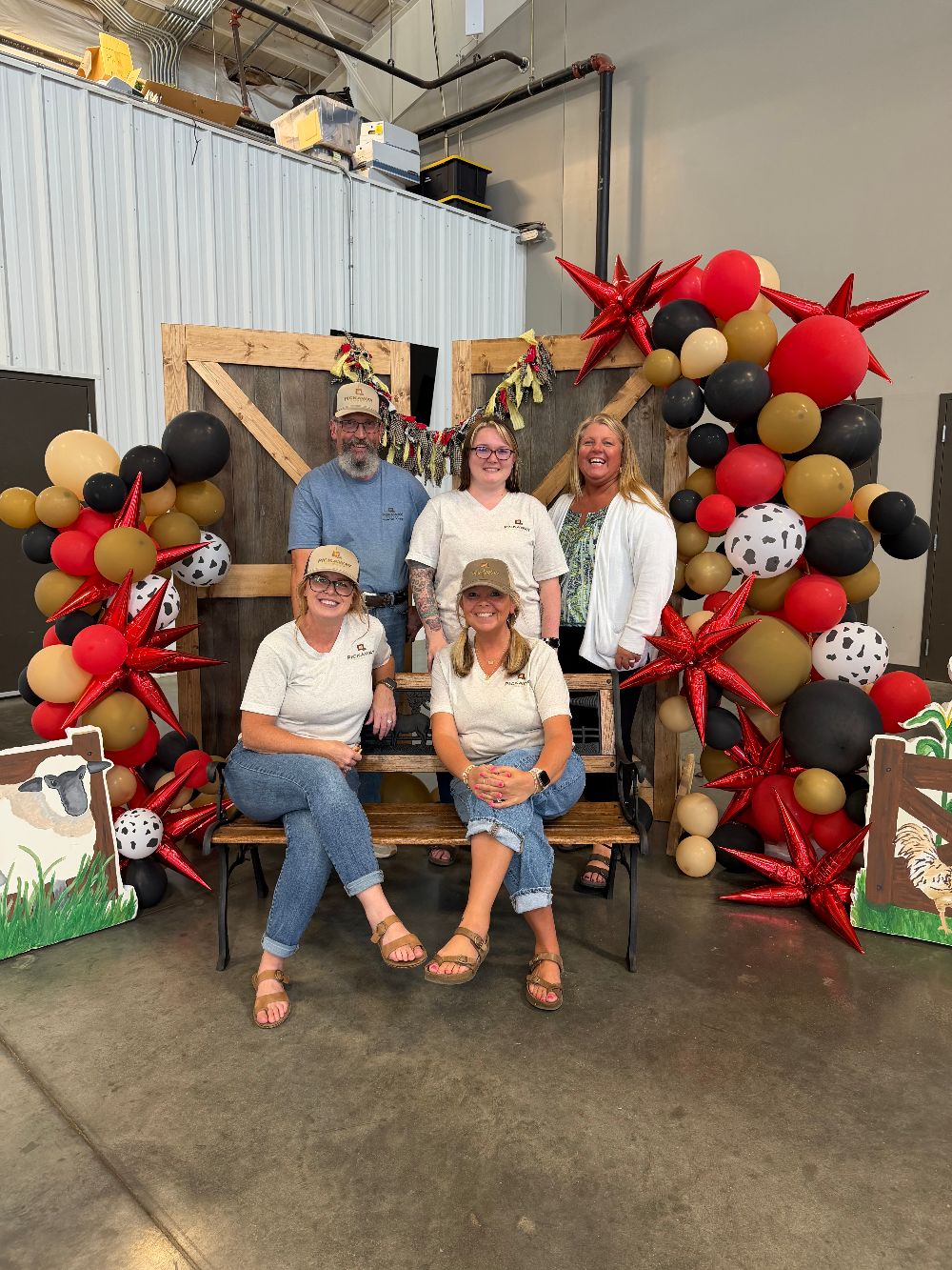 Crystal Fisher with Pickaway County colleagues, posed on and around a wooden bench in front of barn doors decorated with red, black and gold balloons and cow-print balloons.