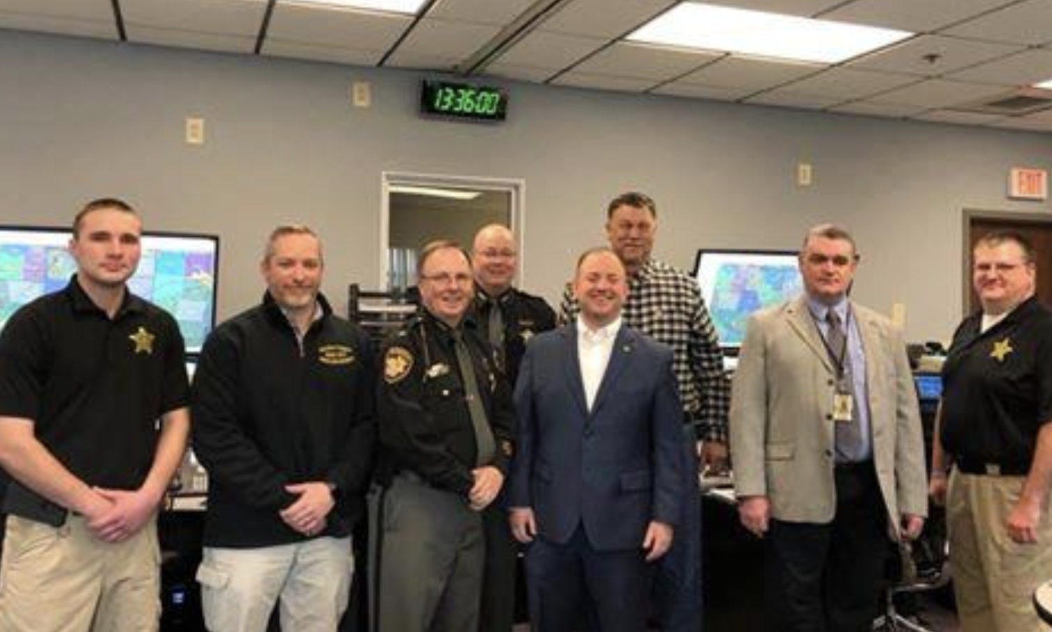 Sheriff's office staff and county officials standing together in the new 9-1-1 dispatch center, with mapping displays and a digital clock behind them