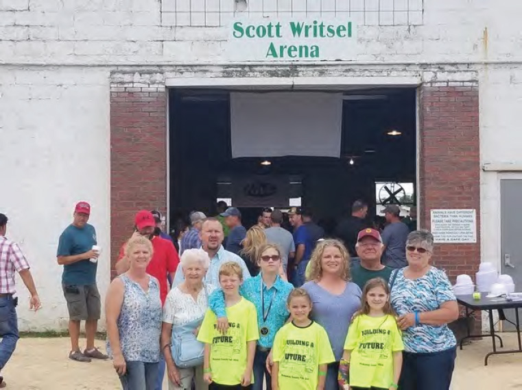 Three generations of a Pickaway County family standing together outside the Scott Writsel Arena at the county fair, the youngest four in matching yellow &ldquo;Building a Future&rdquo; T-shirts, with fairgoers filling the open arena doorway behind them.